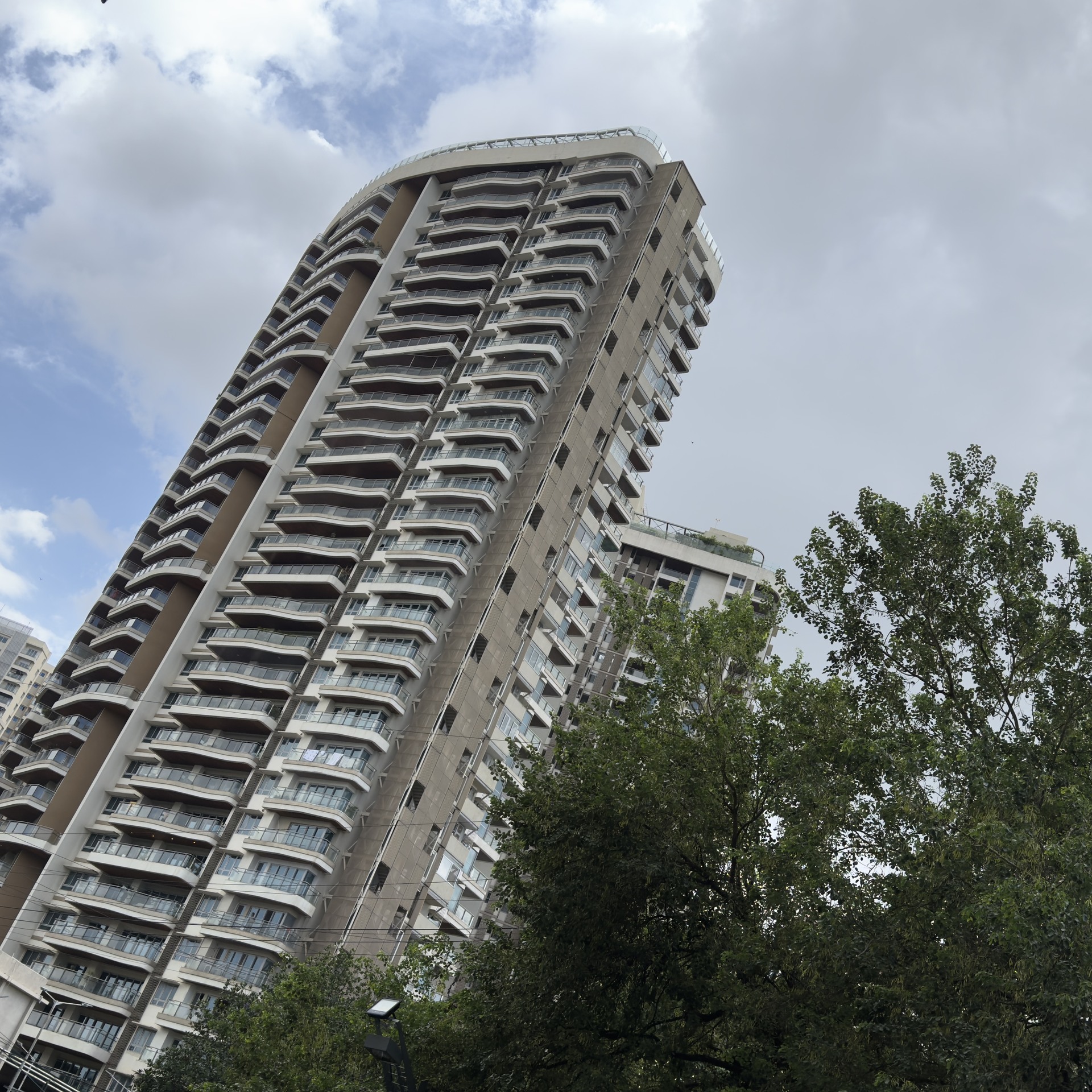 A tall building with balconies, seen from below. Trees cover the lower right side. Clouds cover part of the sky.