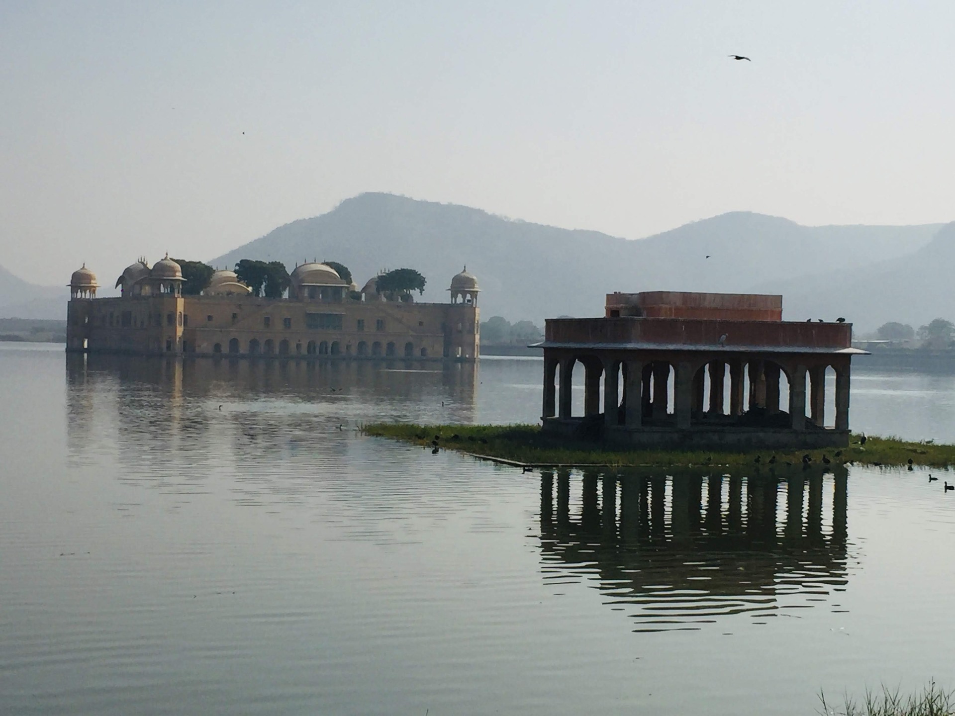 Jal Mahal in a lake in Jaipur. A stone pavilion is in front of the building. Hills are visible behind it.