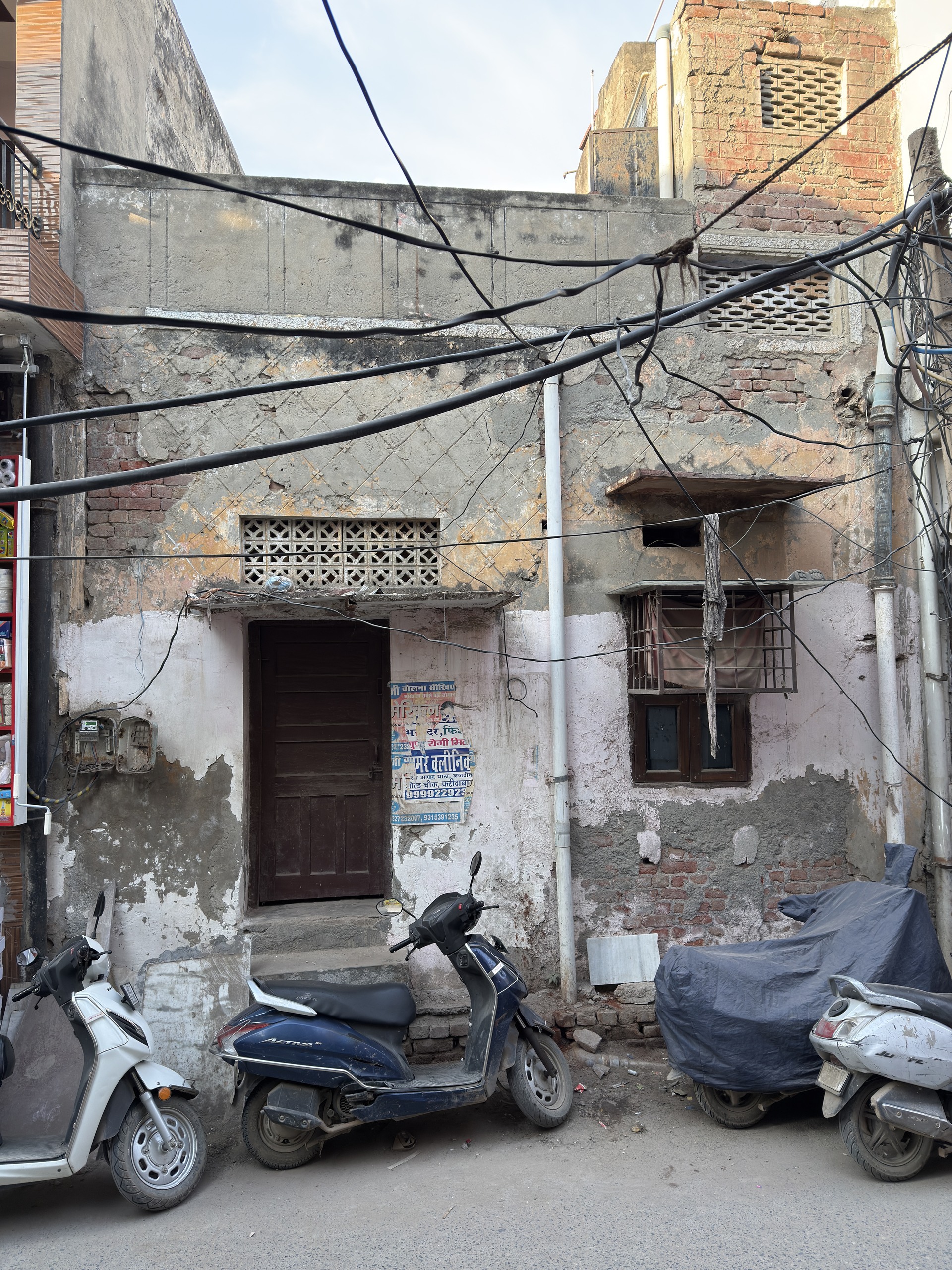 An old building with damaged plaster and exposed bricks. Cables cross the walls. Scooters are parked in front.